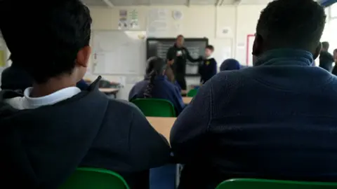 The back of two pupils sitting in plastic chairs as they watch a role play at the front of the class