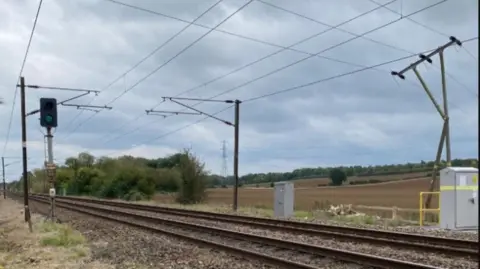 A damaged telegraph pole to the right of the picture shows the wires it carries coming close to the overhead wires above a railway line through the countryside