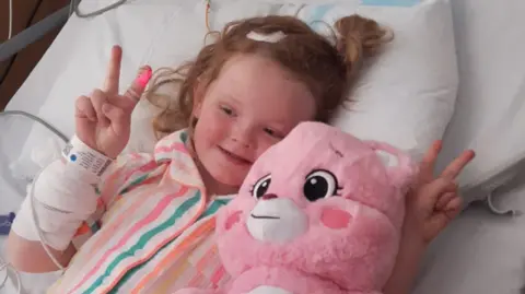 Phoebe is laying on a hospital bed with white sheets. She has ginger hair in a loose ponytail and has a bandage on her arm. She is wearing pink striped pyjamas and is hugging a pink teddy bear.