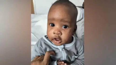 Noah is in a grey baby-suit. He is looking at the camera and appears to be sat on on a bed in a bedroom.