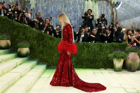 AFP via Getty Images Actress Nicole Kidman arrives for the 2026 Met Gala celebrating "Costume Art" at the Metropolitan Museum of Art, in New York. Kidman wears a long-sleeved beaded column gown in a deep red shade. Red feather ruffles adorn her sleeve cuffs and midriff.