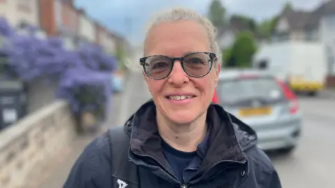 A woman in a navy waterproof jacket stands in a residential street. Homes and cars can be seen blurred behind her. She has tinted glasses and her fair hair is tied back.