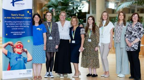 The clinical psychology team at Tom's Trust at Alder Hey Children's hospital - eight women standing side by side next t a Tom's Trust sign 