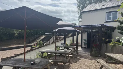 BBC/Chloe Hughes A white building with tiled rood and a veranda over the front door. There are wooden benches outside on a stone area with dark blue umbrellas to shade them from the sun. On the left hand side is a canal lock and some stone steps