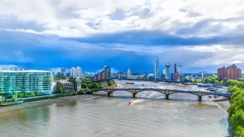 Getty Images A wide shot taking in both sides of the River Thames with high rise modern buildings on both sides and a bridge spanning the river. A dramatic cloud filled sky is at the top of the image. 