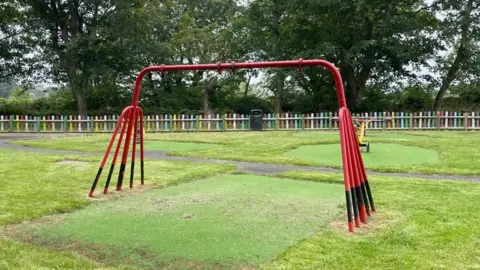 The frame of a red swing set stands in a green play area, enclosed by a multicoloured fence. There are no swings attached to the frame.