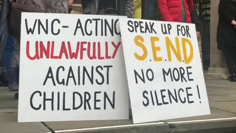 Two placards on the steps of Northampton Guildhall. One reads - 'WNC - ACTING UNLAWFULLY AGAINST CHILDREN', while to its right the other reads 'SPEAK UP FOR SEND NO MORE SILENCE!