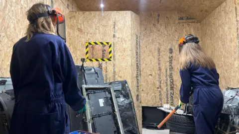 Two women wearing blue overalls in a rage room. The woman on the right has an orange baseball bat in her hand, while the other is looking at her.
