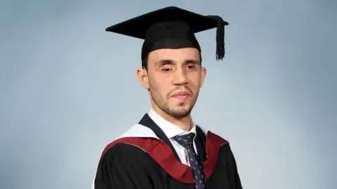 Maher Fattouh in his cap and gown smiling for his graduation photo. He is wearing a black mortarboard, a black robe with red and white accents, a white shirt and a dark blue tie with a white pattern.