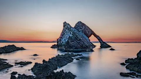 Bow Fiddle Rock, which is very pointy and covered in bird guano, poking out of still sea with sunset behind