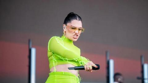 Pop star Jessie J wearing a green outfit and sunglasses on stage pointing down at the crowd while holding a microphone during a performance.