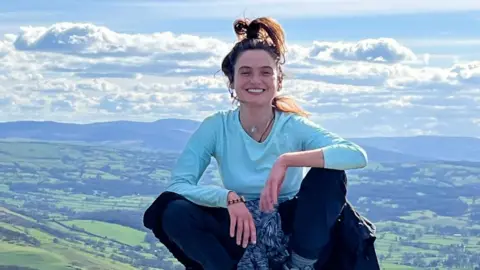 Family photo Eleanor Thompson is pictured sitting on a rock, with the landscape below, wearing a blue top and jeans, with a jumper tied round her waist. Her hair is tied in a high ponytail and she is smiling.