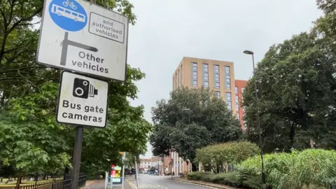 LDRS St Vincent's bus gate, with a sign in the foreground warning of the road restriction and the enforcement camera. A bus stop is situated in the road, between a park and a smaller green area.