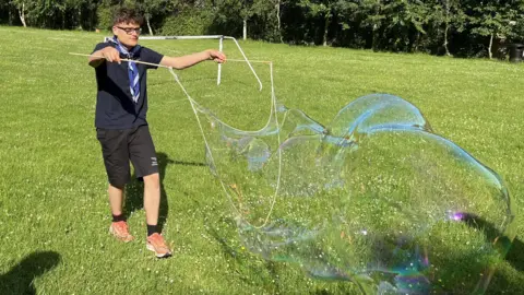 A teenage boy with brown hair and glasses is holding two rods with rope to create a very large bubble on a field