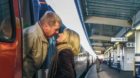 A man on a train and a woman on the platform lean in to each other as if to kiss.