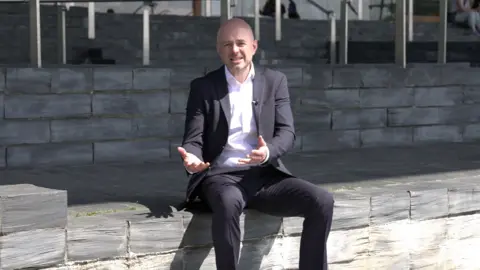 Gareth Lewis sitting on the steps outside the Senedd, wearing a suit and white shirt. He is mid conversation with his hands out in front of him.