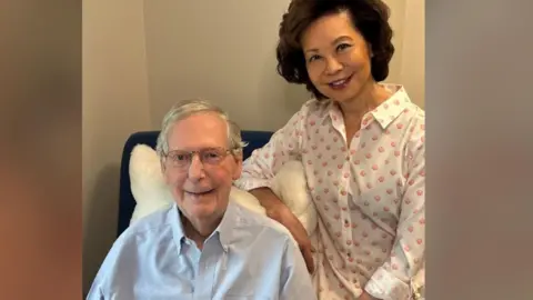 Mitch McConnell sits in a chair and smiles at the camera. His wife Elaine Chao sits next to him and also smiles at the camera. 