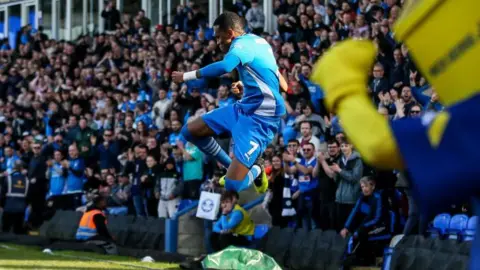 Joe Dent Peterborough United striker Malik Mothersille punches the air as he jumps through the air in front of a stand of Peterborough United fans.