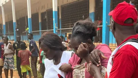 Ulrich Tobet A man and a woman holding another woman who looks visibly traumatised outside the school