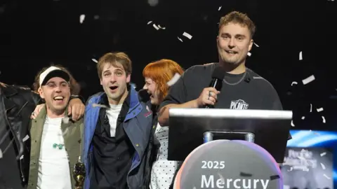 PA Media Sam Fender at the podium after winning the Mercury Prize in Newcastle. He is holding a microphone and smiling. Confetti is raining down around him and his band, who are standing behind him with their arms around each other.