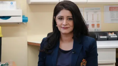 Emma Baugh/ BBC Dr Shabina Asad Qayyum smiles at the camera while sitting in an office. She is wearing a navy blue top and a navy blue blazer with an orange logo. She has long black hair. 