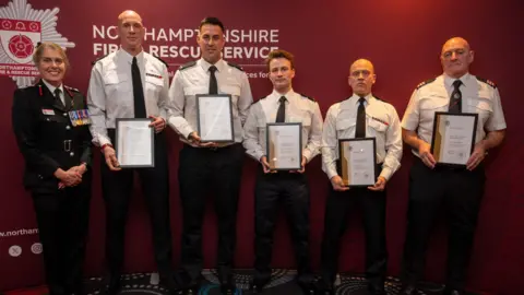 Chief fire officer Nikki Watson, dressed in black uniform with lots of different coloured medals, smiles at the camera as she stands next to five men, all in white shirts and black ties, trousers and shoes. They are holding award certificates. 