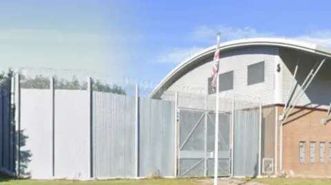 The white outside of Deerbolt prison, which has a domed roof. There is an unfurled union jack flag on a pole outside the gate. 