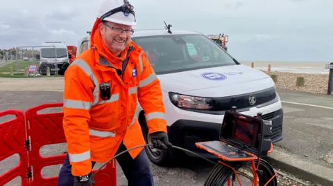 A man in an orange hi-viz jacket looking at a camera down a manhole cover.