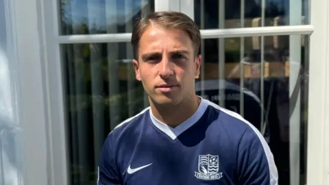 Alexis Cassar looking straight at the camera with a neutral expression. He is standing outside, in front of a window, and is wearing a navy blue football shirt with a white Nike tick on the left and a white Southend United badge.