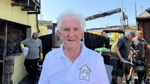 Len Curtis, with white hair and a white shirt, poses for a photo with workmen repairing the home in the background
