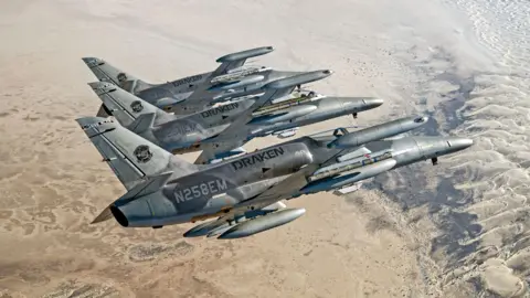 Draken Europe Draken's Alca L-159E "Honey Badger" aircraft flying in formation. Sand covers the ground below them. The three planes are lined up and flying to the right. They are tilted away from the camera as if turning.