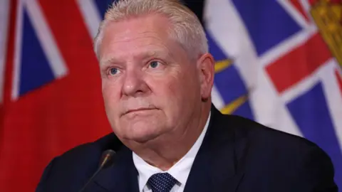 Doug Ford seated before a microphone at a press conference. His expression is neutral. 