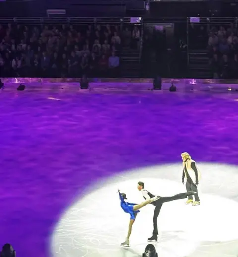 Suzanne Otterson-MacDonald Lewis and Isla perform on the ice, both caught mid-arabesque. Isla is in a blue skating dress, Lewis in a black waistcoat suit. A coach guides them through their training.
