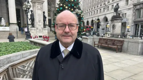 Chris Hayward is pictured in front of the Royal Exchange, an older white-stoned building which has statues, a Christmas tree and poppy wreaths in the background