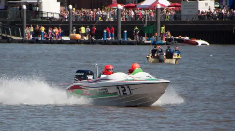 A boat, on the Great Ouse river. The boat is white with green and red detailing, and the number 121 displayed on the side. The driver is wearing a bright red helmet. In the background, people line the top of the river bank, and the dock below it.