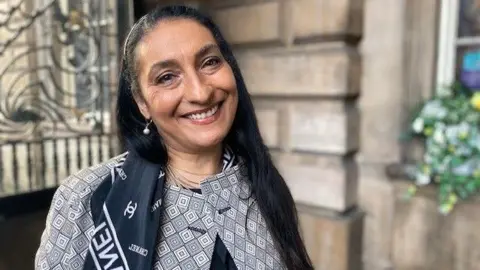 BBC/Claire Hamilton Linda Priddie smiles for the camera, she is wearing a navy blue Chanel scarf and a blazer with a black, white and grey geometric pattern. She has long dark hair and is standing outside Liverpool Town Hall, whose stonework can be seen in the background.