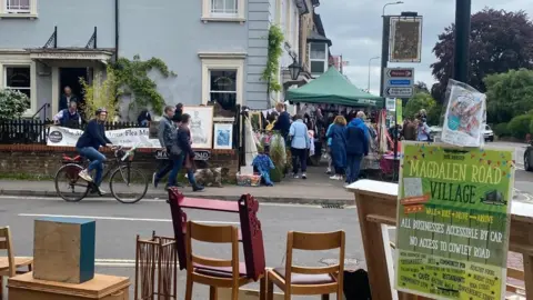 Emily Kerr Magdalen Road Food Festival - an image taken looking down the stalls on the road from behind various types of furniture at another stall. Several people and a dog can be seen in the street. There is bunting hung around one stall under a green marquee