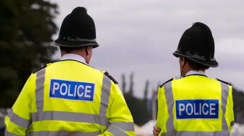 Two police officers stood outside, with their backs to the camera. They are both wearing police hats and high vis police jackets