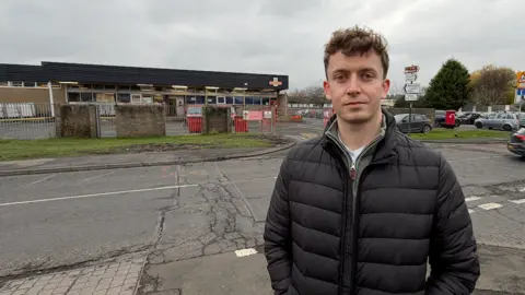BBC/JIM SCOTT Councillor Brandon Feeley is standing outside a Royal Mail sorting office. He has short curly brown hair and wears a black padded jacket. Across the road behind him is the single storey delivery office. It has dark horizontal wood panelling at the top, with a Royal Mail logo on the extreme right hand corner. Below are lots of windows and a wall with metal fencing in front.