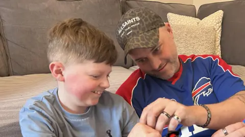 Kate Bradbrook/BBC On the left is a young boy in a grey T-shirt smiling as he plays with Lego with his dad, right, who is wearing a baseball cap and blue and red sports top.