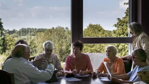 Victor Steadman Six retirement village residents sit around a table in front of a big window. They have pens and paper in their hands. One woman stands behind one of the resident's chairs