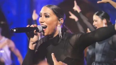 Aston Band Marina Berry-Dealey with slicked back black hair pulled into a ponytail performing at the Royal Variety Show after party. She is singing with a microphone held in her right hand and up to her mouth and her left arm is outstretched wide. Behind her are other singers in black, pointing with their upraised right hands.