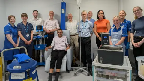 Sheffield Teaching Hospitals NHS Foundation Trust A team of doctors and nurses are standing behind a patient in a chair and posing for a photograph. They are surrounded by various bits of medical equipment, including a drip stand and blood pressure machine.