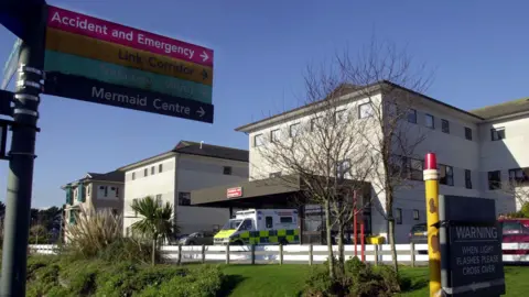 A general view of the Royal Cornwall Hospital in Truro on a sunny day. In the foreground is a sign pointing to Accident and Emergency, Link Corridors, Trelawny Wing and Mermaid Centre. There is an ambulance parked outside the main entrance to the building. To the right is a sign which reads: Warning when light flashes please cross over. 