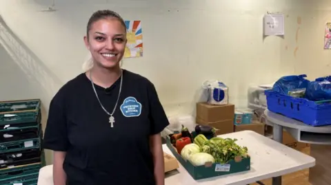 Amelia - a young woman with tied back hair wearing a black t-shirt with the organisation's logo on in and an ankh necklace. She is standing in front of stacks of boxes containing food