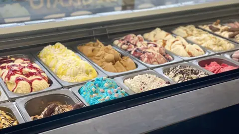 Nick Price A row of colourful ice cream flavours is displayed in metal trays inside a glass counter.
