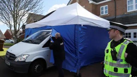 An officer pulls part of a blue and white police tent which has been placed over a white van outside a brick house. On the right of the picture a police officer in a high viz vest looks on.