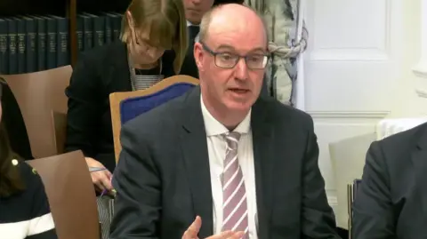 Neil Guckian giving evidence at Stormont's Health Committee. He is bald and wearing glasses and black blazer, white shirt and pink and white tie. A man and a woman are sitting next to him.