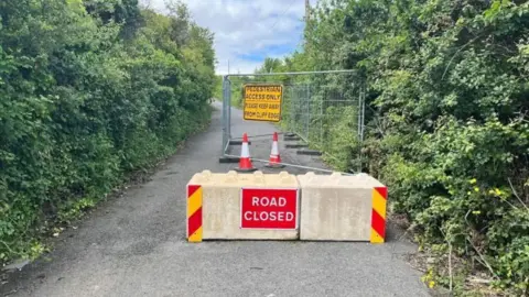 BBC A country road is closed with concreate barriers with a red 'road closed' sign on it. There is also metal cage fencing behind the barrier with a yellow sign that says 'Pedestrian access only, please keep away from cliff edge'.