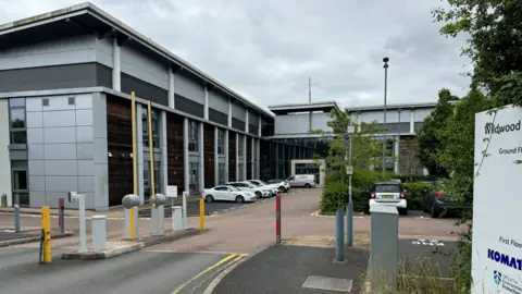 A picture showing a car parking barrier, and a large office block - parked outside are several cars. 